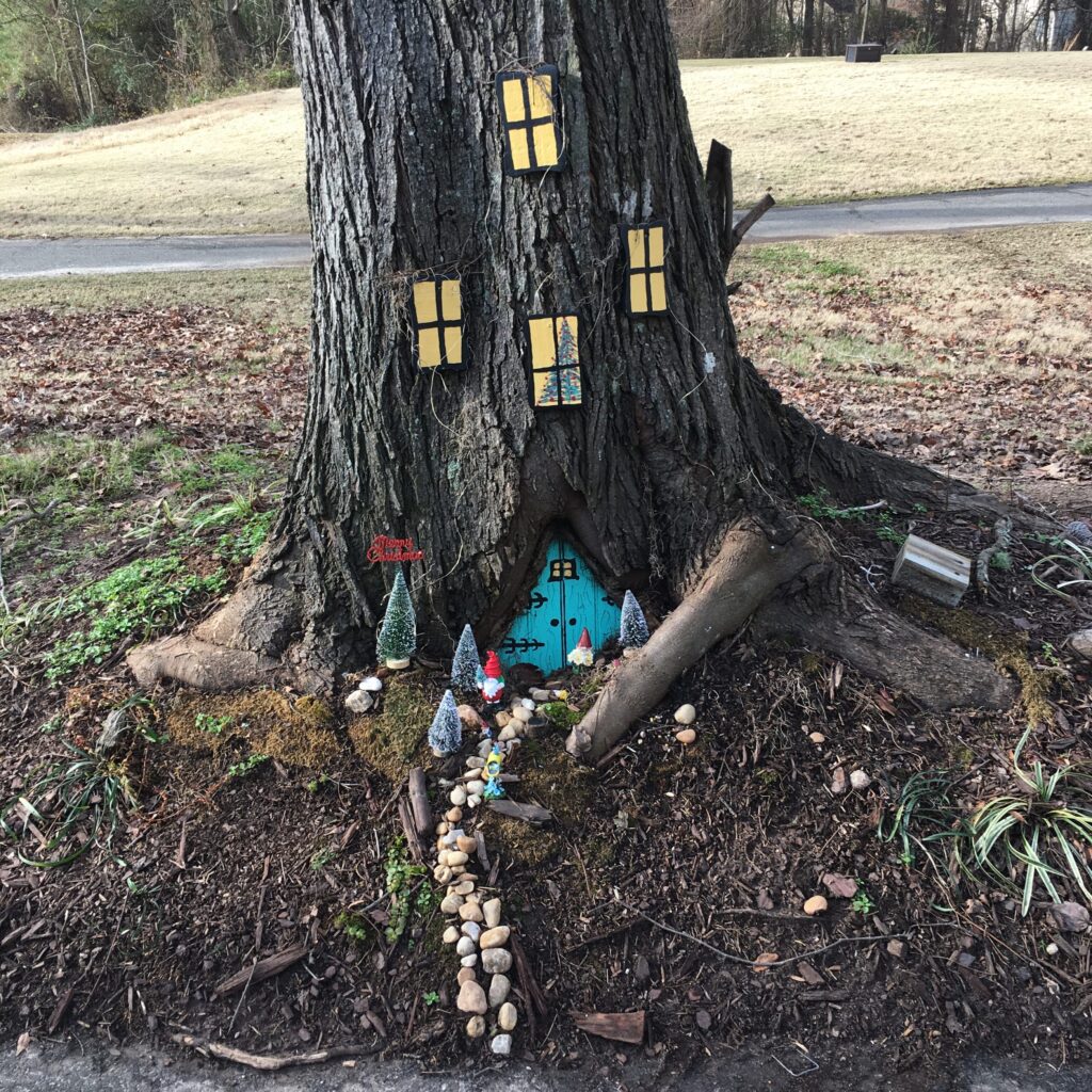Tree trunk decorated with windows and a door at the base, a row of mushrooms lead to the door like a paved walkway