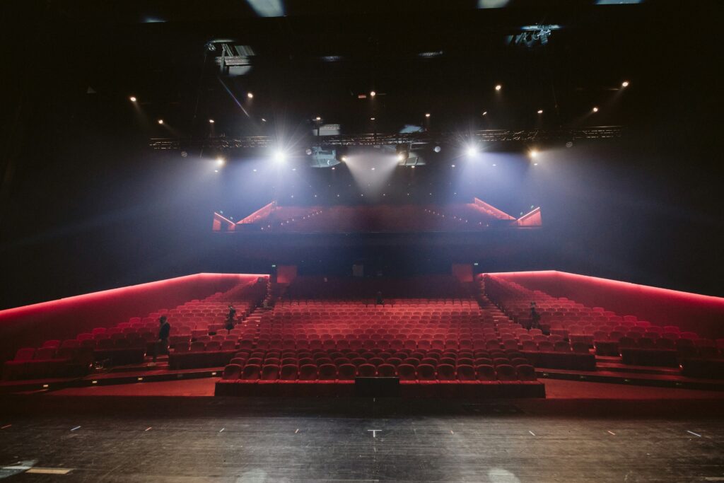 Photo of a theater as seen from the stage, part of the stage at the bottom and rows of red upholstered seats rising up to the back/middle of the photo, with the balcony seating at the back/top along with the lighting along the ceiling