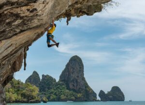 Photo of a person hanging from a nearly horizontal cliff face above the ocean, other distant mountains in the background